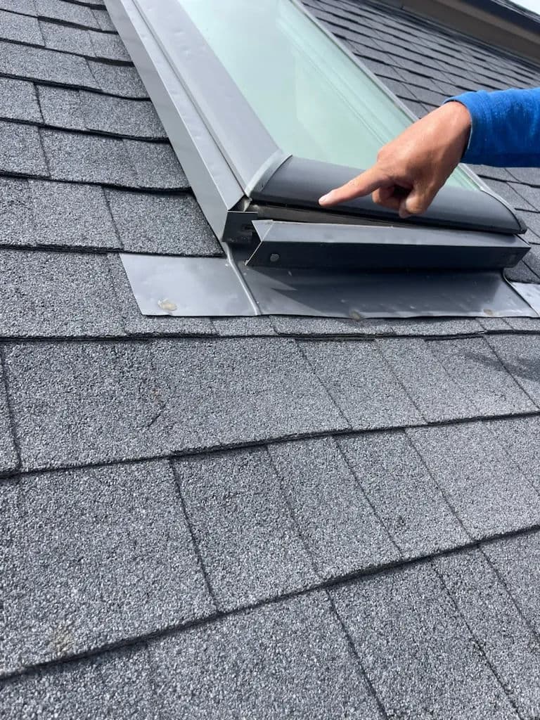 Hand pointing at a skylight installation on a textured black shingle roof.