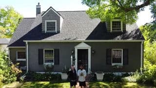 Two people stand in front of a charming gray house with a sloped roof and green landscaping.