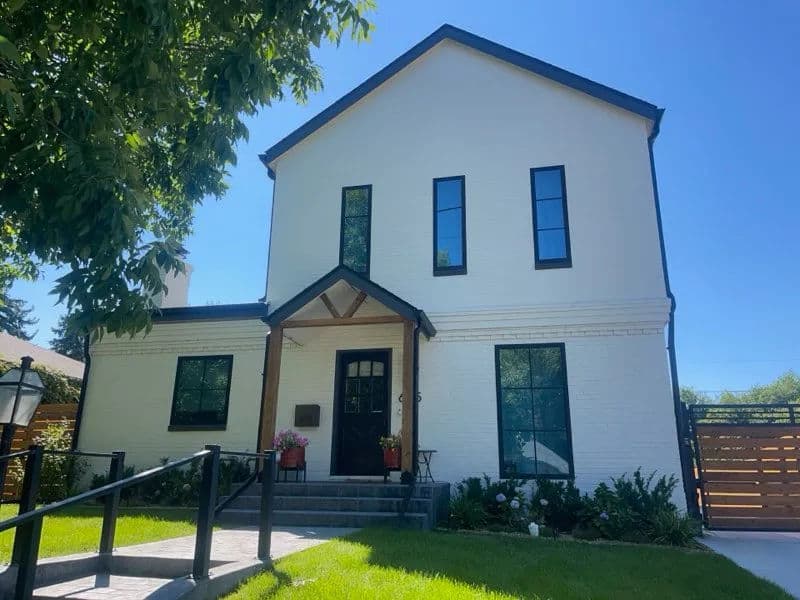 Modern white home with black trim, large windows, and landscaped front yard. Bright sunny day.
